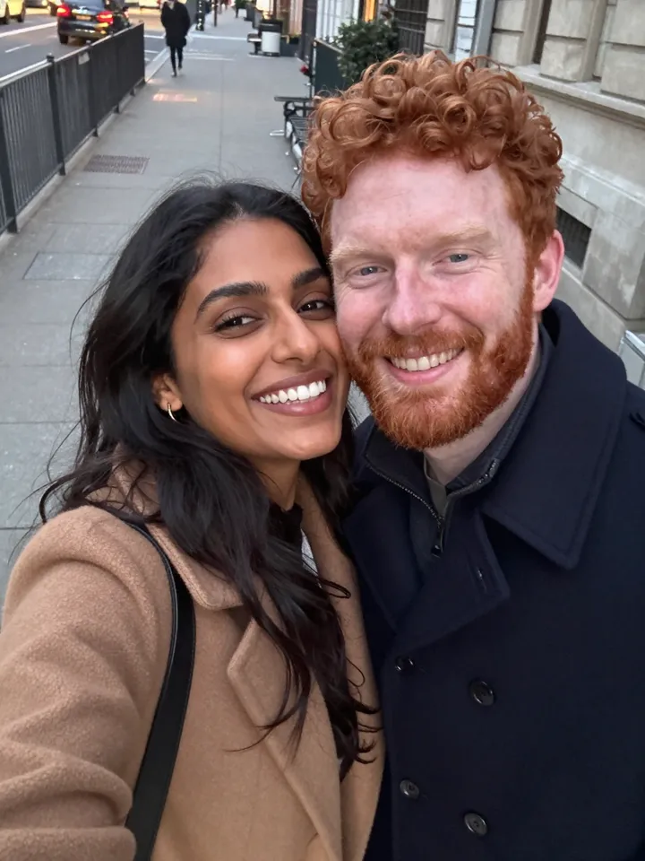 A couple in their thirties taking a selfie on a city sidewalk at dusk, cheeks pressed together
