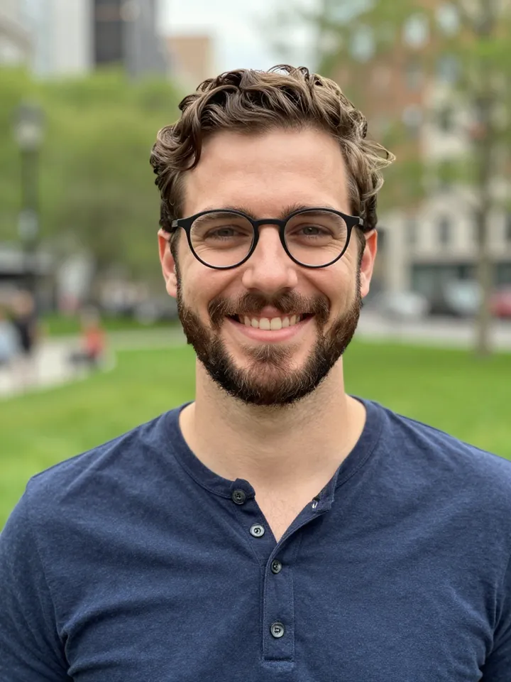 Portrait photo of a smiling man in his thirties with wavy brown hair, a short beard and black-framed glasses in a park