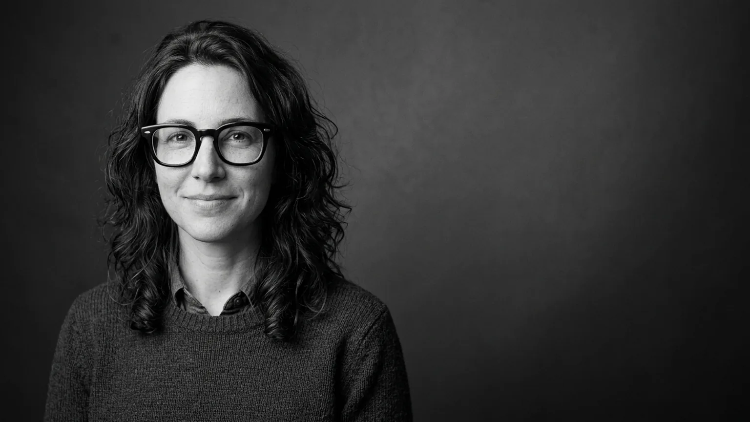 Black-and-white studio half portrait of a fictional woman with wavy hair and thick black glasses, dark gradient backdrop with space on the right