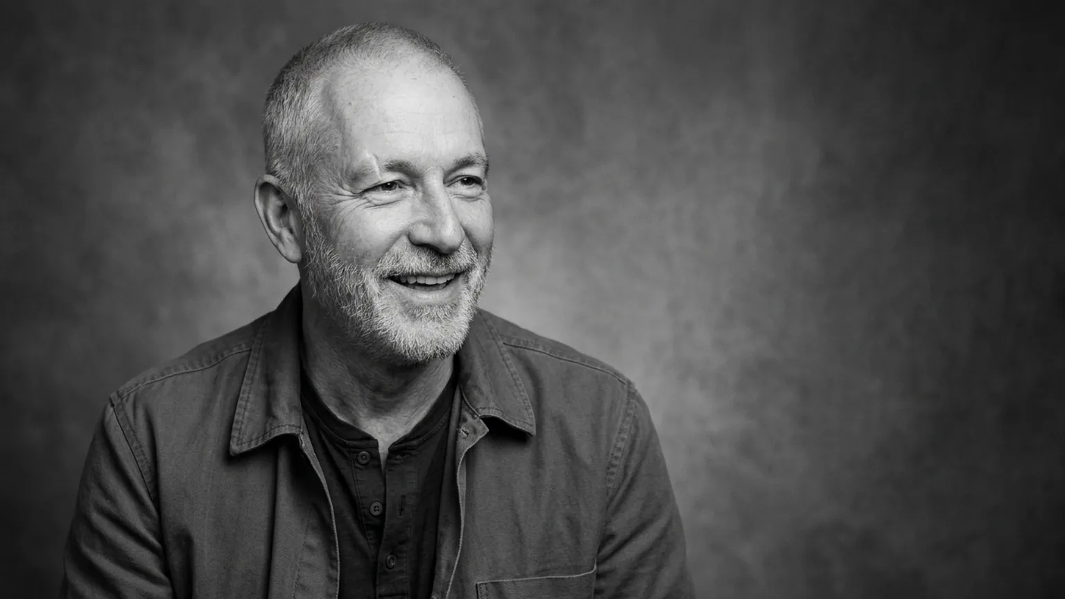 Black-and-white studio portrait of a fictional older man with a short silver beard laughing, soft light from the left